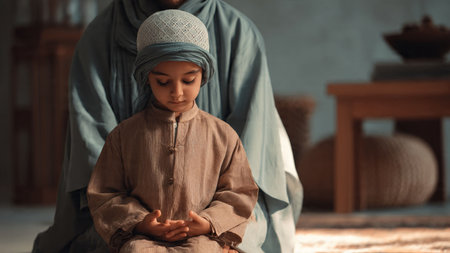 A young Muslim boy sits in prayer, hands clasped, wearing traditional attire, with an adult figure in similar dress behind him, conveying a sense of spiritual orientation and family bonding.の写真素材