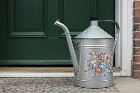A silver metal watering can with floral design sits on a tiled front porchの写真素材
