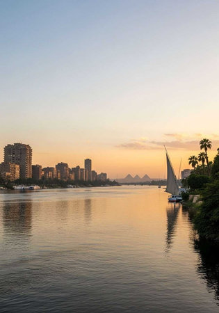 A serene cityscape at sunset with a sailboat on the calm waters of a riverの写真素材