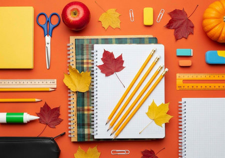 Overhead view of notebooks, pencils, scissors, and autumn leaves arranged on vibrant orange surfaceの写真素材