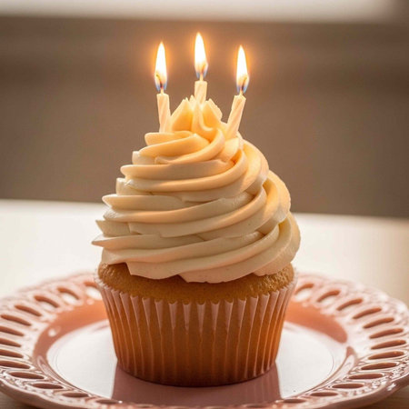 A cupcake with white frosting and three lit candles on top sits on a pink plateの写真素材