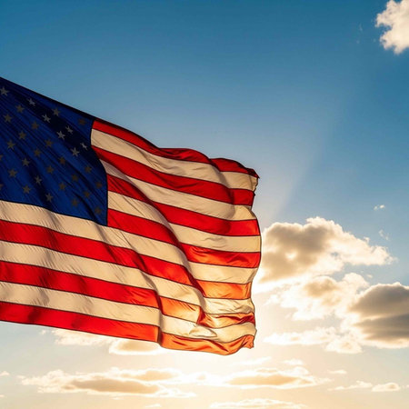 A large American flag flies in the wind against a clear blue sky with white clouds and sunshine.の写真素材