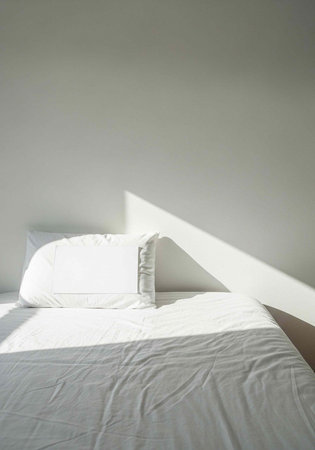 A neatly made bed with white sheets and pillows, illuminated by sunlight casting a diagonal shadow on the wallの写真素材
