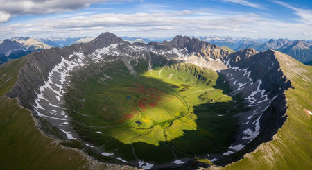 Panoramic landscape of rugged mountains, snow patches, and lush green valley under a partly cloudy blue skyの写真素材