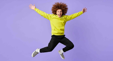 Excited curly haired young woman in yellow sweatshirt jumping and looking at camera isolated over violet background