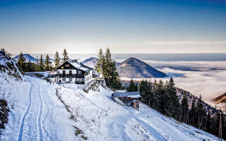 A small house on a snowy mountainside next to narrow road. View of Alps above cloudsの写真素材