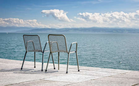 Two empty metallic chairs on the shore of a large lake. Shot from behind the chairsの写真素材