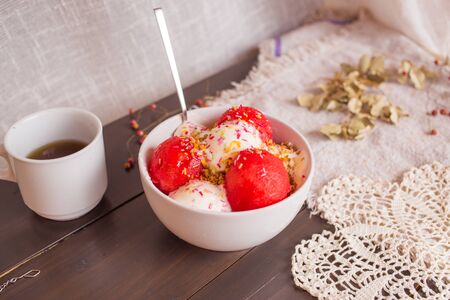 Delicious dessert with vanilla ice cream, watermelon and sprinkles in white cup with tea on dark wooden table covered by napkin and knitted doilyの写真素材