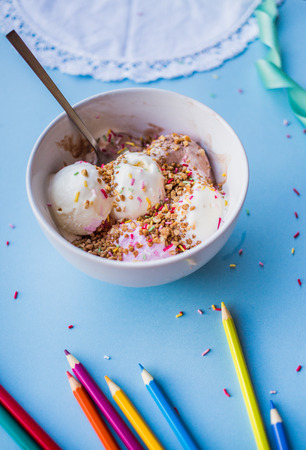 Delicious dessert with vanilla and strawberry ice cream and sprinkles in white cup on blue background with color pencils and white napkinの写真素材