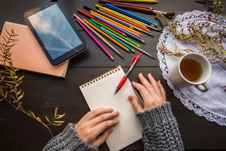 Hands with notebook with squared paper at the dark wooden table with cup of tea, color pencils, pad, orange book and dry plantsの写真素材