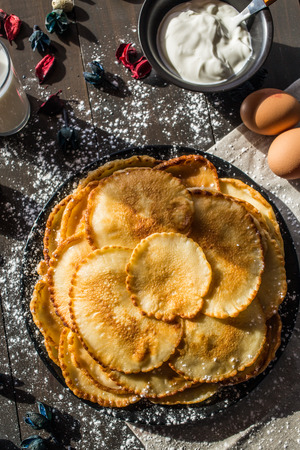 Sweet delicious russian pancakes on a black plate, sprinkled with powdered sugar, with a glass of milk and a cup of sour cream on a table of dark wood, covered with beige napkinの写真素材