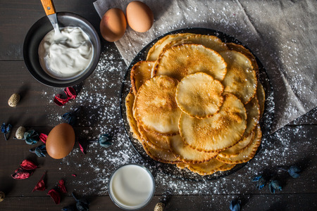 Sweet delicious russian pancakes on a black plate, sprinkled with powdered sugar, with a glass of milk and a cup of sour cream on a table of dark wood, covered with beige napkinの写真素材