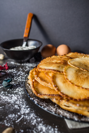 Sweet delicious russian pancakes on a black plate, sprinkled with powdered sugar, with a glass of milk and a cup of sour cream on a table of dark wood, covered with beige napkinの写真素材