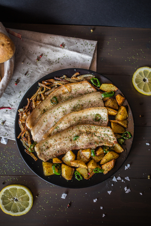 Delicious fried pork with bits of potato on a black plate on a table of dark wood, covered with beige cloth, with slices of lemon, salt and a basket of breadの写真素材