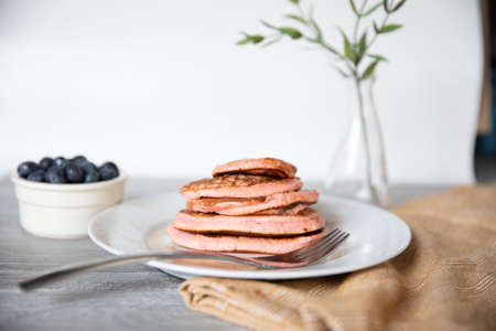 Pancakes stacked with syrup and blueberries and a napkin and vase with greenery. Fork and grey wood ...