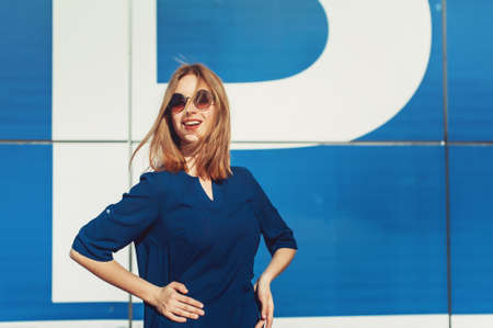 Cute fashionable business woman in dress outdoors. Stylish blonde woman in fashionable blue dress walking on the summer city street near the blue wall of business centre building. の写真素材