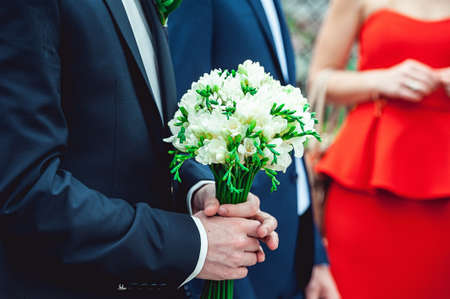 fiance in a dark blue suit holds a wedding bouquet made of white flowersの写真素材