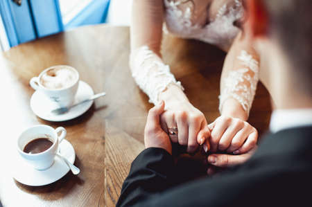 the bride and groom drinking coffee in a cafe at the round wooden table. gently holding each others handsの写真素材