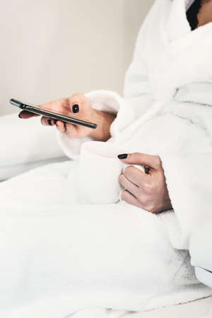 Attractive young girl in bathrobe using smart phone, holding a white cupの写真素材