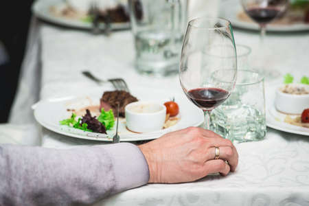 Mans hand holds a glass of wine. fresh grilled bbq roast beef steak and sauce on a white plate with green leaf of salad. soup sauce small jug glass served on a table in a restaurantの写真素材