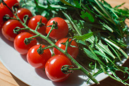 Whole red tomato cherry and fresh ruby leaves composition on a white plate on the wooden tableの写真素材