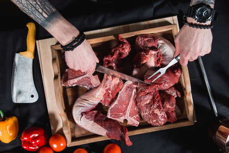 raw meat different kinds on a board on a table. Knife and fork in hands of the chef. Cook offers steaks to choose from. Close-up in the kitchenの写真素材