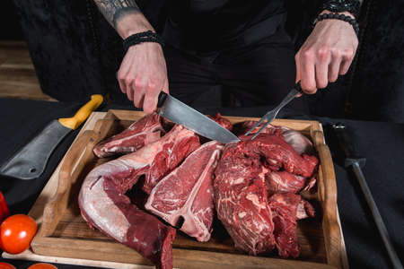 raw meat different kinds on a board on a table. Knife and fork in hands of the chef. Cook offers steaks to choose from. Close-up in the kitchenの写真素材
