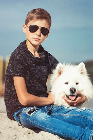 Happy 11 year old boy hugging his dog breed Samoyed at the seashore against a blue sky close up. rest and have fun on vacation, play in the sandの写真素材