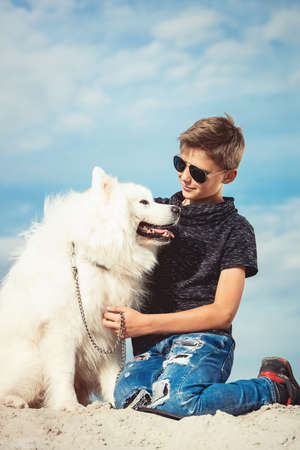 Happy 11 year old boy playing with his dog breed Samoyed at the seashore against a blue sky close up. Best friends rest and have fun on vacation, play in the sandの写真素材