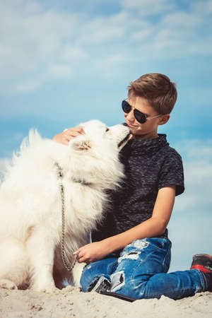 Happy 11 year old boy playing with his dog breed Samoyed at the seashore against a blue sky close up. Best friends rest and have fun on vacation, play in the sandの写真素材