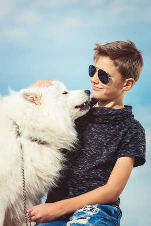 Happy 11 year old boy playing with his dog breed Samoyed at the seashore against a blue sky close up. Best friends rest and have fun on vacation, play in the sandの写真素材