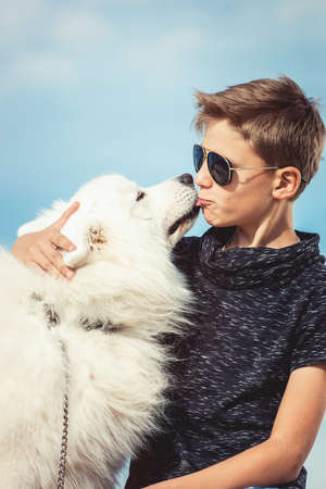 Happy 11 year old boy playing with his dog breed Samoyed at the seashore against a blue sky close up. Best friends rest and have fun on vacation, play in the sandの写真素材