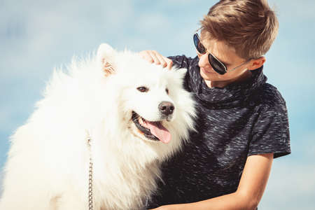 Happy 11 year old boy playing with his dog breed Samoyed at the seashore against a blue sky close up. Best friends rest and have fun on vacation, play in the sandの写真素材