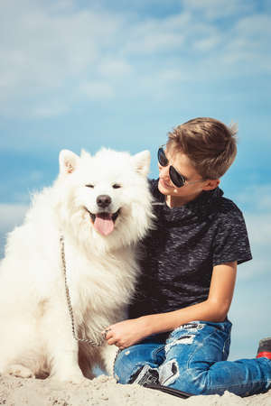 Happy 11 year old boy playing with his dog breed Samoyed at the seashore against a blue sky close up. Best friends rest and have fun on vacation, play in the sandの写真素材