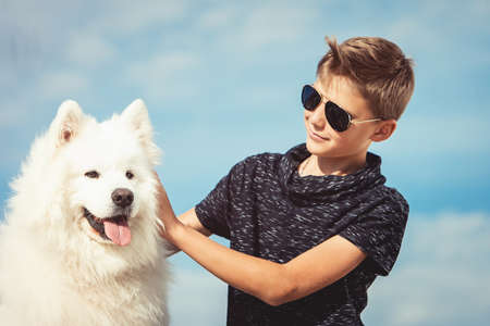 Happy 11 year old boy playing with his dog breed Samoyed at the seashore against a blue sky close up. Best friends rest and have fun on vacation, play in the sandの写真素材