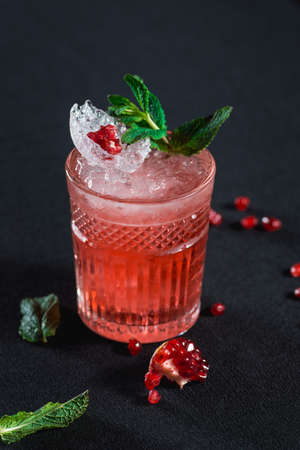 Cold cocktail with ice and garnet seeds, piece of pomegranate behind on dark backgroundの写真素材