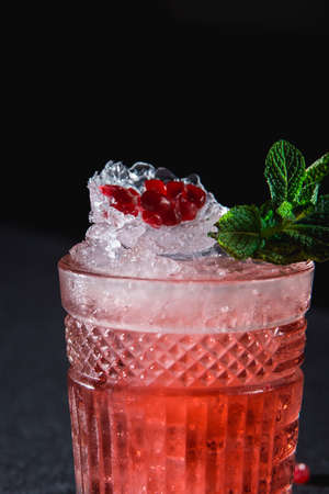 Cold cocktail with ice and garnet seeds, piece of pomegranate behind on dark backgroundの写真素材