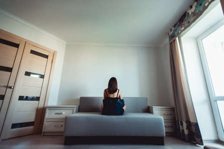 Loneliness. Alienation. Girl in an empty room sits back on a couch and looks in the wallの写真素材