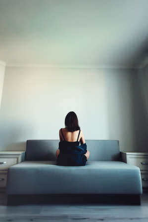 Loneliness. Alienation. Girl in an empty room sits back on a couch and looks in the wallの写真素材