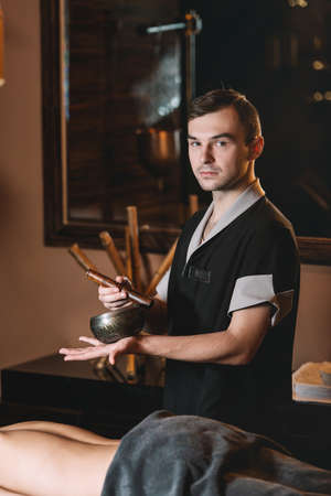 Specialist holding in hands Brass Hammer Singing Bowl. Young charming girl on a panchakarma procedure laying on a massage table. beautiful woman spending time at modern spa cabinet relaxing. Soft yellow light.の写真素材