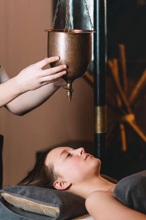 Specialist making shirodhara massage form of ayurveda therapy. Young charming girl on a panchakarma procedure laying on a massage table. beautiful woman spending time at modern spa cabinet relaxing. Soft yellow light.の写真素材