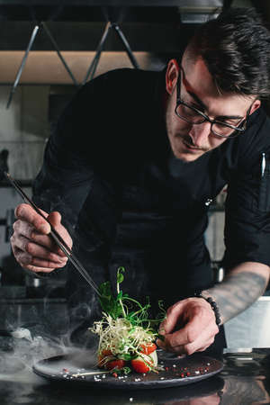 Chef finishing healthy salad on a black plate with tweezers. almost ready to serve it on a table. Working with smokeの写真素材