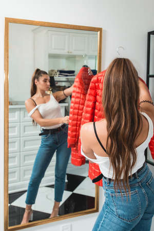 Young woman in a blu jeance white t-shirt trying on red down coat in a luxury boutique. Standing in a front of a mirror in a white roomの写真素材