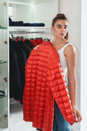 Young woman in a blu jeance white t-shirt trying on red down coat in a luxury boutique. White roomの写真素材