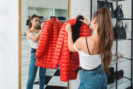 Young woman in a blu jeance white t-shirt trying on red down coat in a luxury boutique. Standing in...