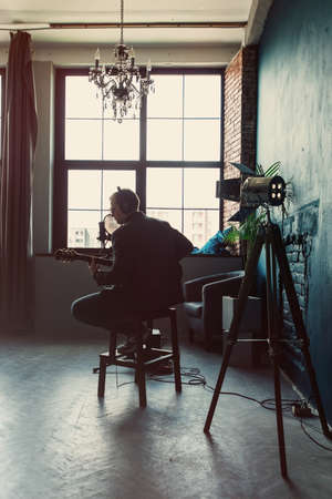 Close up of a man singer in a headphones with a guitar recording a track in a home studio. Man wearing sunglasses, jeans, black shirt and a jacket. Look from back, window on a bakgroundの写真素材