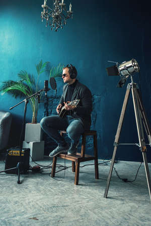 Close up of a man singer in a headphones with a guitar recording a track in a home studio. Man wearing sunglasses, jeans, black shirt and a jacket. side viewの写真素材