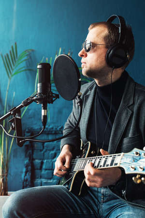 Close up of a man singer in a headphones with a guitar recording a track in a home studio. Man wearing sunglasses, jeans, black shirt and a jacket. side viewの写真素材