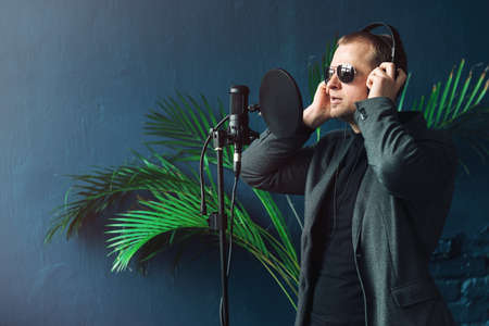 Close up of a man singer in a headphones recording a song in a home studio. Man wearing sunglasses, black shirt and a jacket. side viewの写真素材
