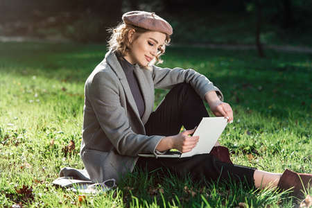 Work outdoors benefits. Education technology and internet concept. Smiling girl in a beret work with laptop in park sitting on grass. Natural environment office.の写真素材